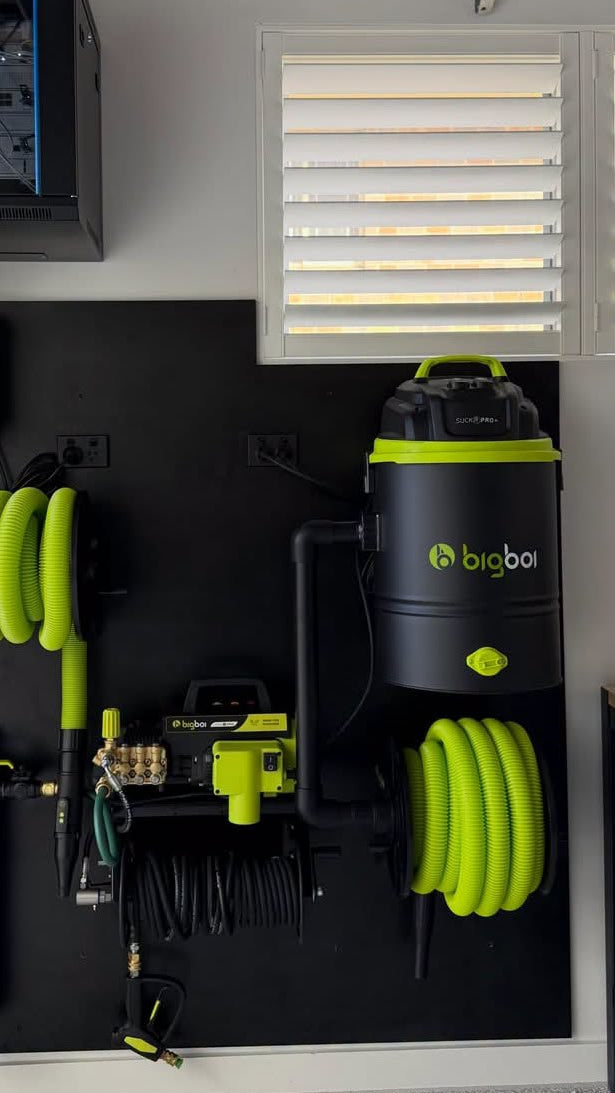 A wall-mounted black garage organizer panel holding multiple cleaning tools, including two large coiled neon green hoses, a black canister with a visible BigBot logo, filter canisters, a yellow pressure washer attachment, and a red tool hanging on the left.